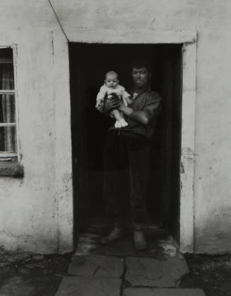 Standing in a darkened doorway, a man covered in coal dust holds an infant in light-colored clothing