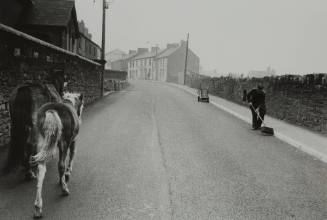 Walled village street with two horses on the left of image, a man sweeping gutter on the right