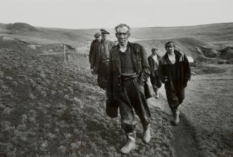 Men with soot on their faces and wearing baggy, dark, work clothes walk on path on a steep hillside