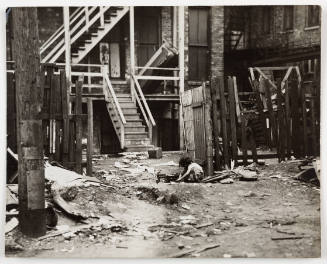 Black-and-white photo of back of apartment building and child in foreground playing among debris