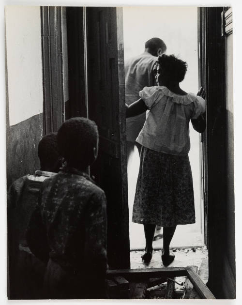 Photo of two adults and two children with dark skin tone seen from behind, walking through doorway