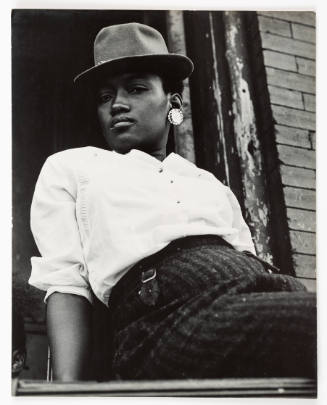 Black and white photograph of dark-skinned adult in fedora and button-up shirt looking at camera