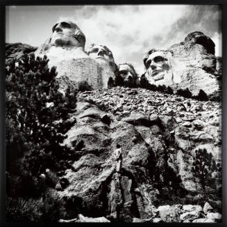 Black-and-white landscape with human faces carved in rock with small figure at center of the image 