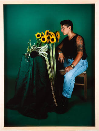 A young, light-skinned adult with tattooed arm sits in a green-toned room facing standing sunflowers