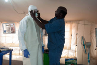 Inside a tent, a man adjusts goggles on a person in full-body personal protective equipment