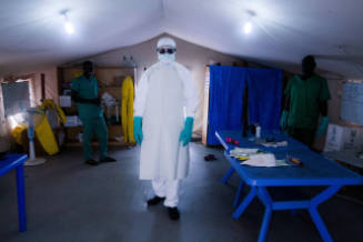 Inside medical tent, person in full-body personal protective equipment and two people wearing scrubs
