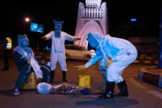 Outdoors and at night, three figures in masks and protective equipment surround a body on the ground