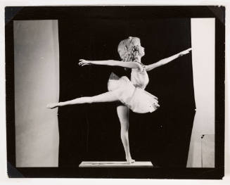 Doll-like figure wearing tutu stands in ballerina pose with arms outstretched and leg raised behind