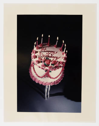 Color photograph of pink-trimmed birthday cake with lit candles and human legs