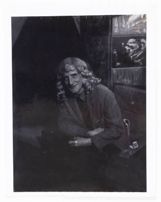 Dimly lit photograph of an older adult with shoulder-length hair leaning forward in chair 
