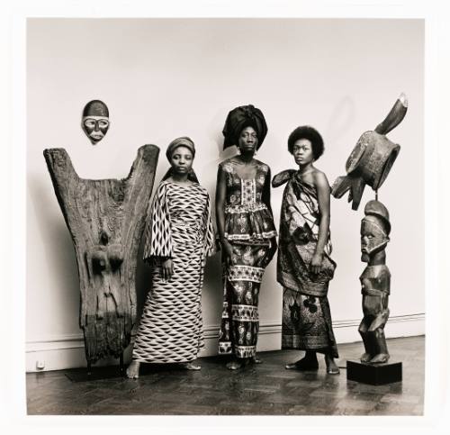 Photo of three dark-skinned women standing between two sculptures and wearing patterned dresses 