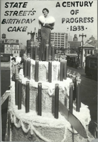 A young woman stands atop a grand birthday cake parade float in evening attire with photo caption
