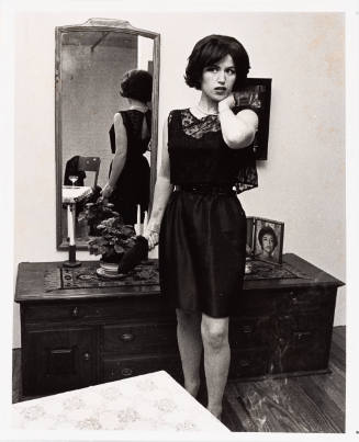 A fair-skinned woman stands in front of a mirror in a black dress, looking to the left of the camera