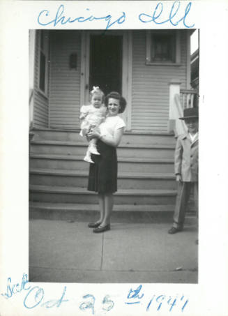 Family snapshot of woman holding baby, near house front steps, with young boy in formal hat at right