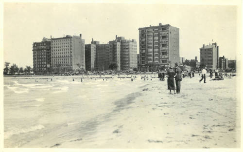 Beach city scape, shore and clothed figures in foreground, bathers and and buildings in background