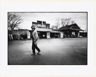 Black-and-white photo of a man walking to the left, past remnants of gas station or general store