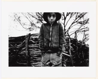 Black-and-white photo of a child in front of a stick fence looking into camera with hands in pockets