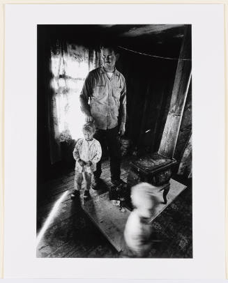 Black-and-white photo of middle-aged man and two children in spare, wooden room with iron stove