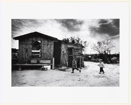 Two children running in foreground with a run-down house, barren trees, and vast sky behind them