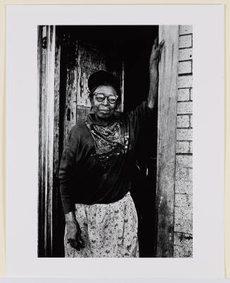 Older African-American woman with glasses standing in a doorway facing viewer with arm raised