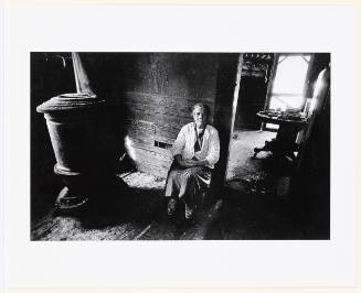 Black-and-white photo of older woman with direct gaze sitting in spare, wooden room with iron stove
