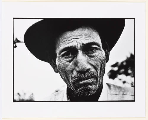 Black-and-white portrait of an older man in hat with medium skin tone and wrinkles gazing at camera