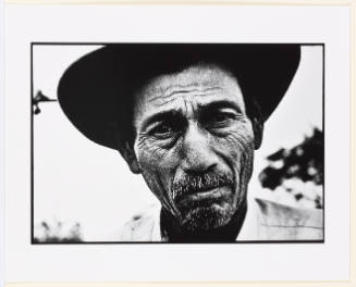 Black-and-white portrait of an older man in hat with medium skin tone and wrinkles gazing at camera