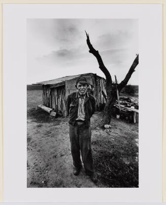 Portrait of older male-presenting figure in front of a corrugated metal structure and a forked tree