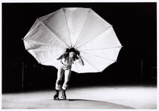 Black-and-white photo of Robert Rauschenberg on roller skaters carrying an umbrella-like stage prop