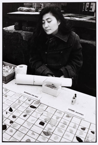 Photograph of Yoko Ono seated at a table and looking at shards of glass placed on a grid 