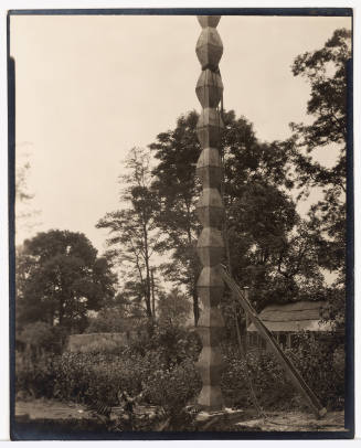 A ladder leans against a tall metal column sculpture in a garden with small building in background