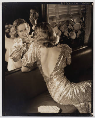 Sepia-toned photograph of a woman in a fancy dress seen from behind and smiling in a mirror