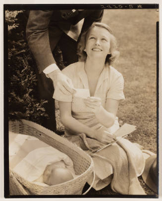 Smiling light-skinned woman sitting on grass next to a baby showing papers to a man bending over her