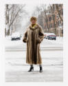 An older woman with medium skin tone wearing a fur coat standing on a sidewalk dusted with snow