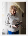 An older, light-skinned person wearing makeup, holding a toy plane, standing in front of a window