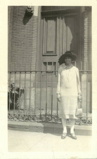 A Black woman in a dress and brimmed hat standing in front of an iron fence of a brick building