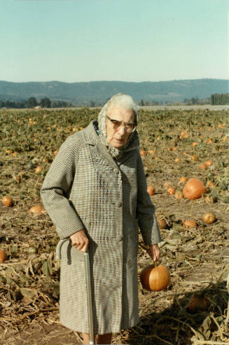 Older white woman wearing a headscarf and coat holding a pumpkin in a pumpkin field