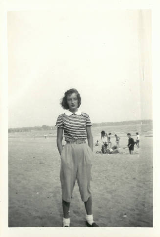 Teenage girl standing with hands in pockets of pants on a beach with people in the background