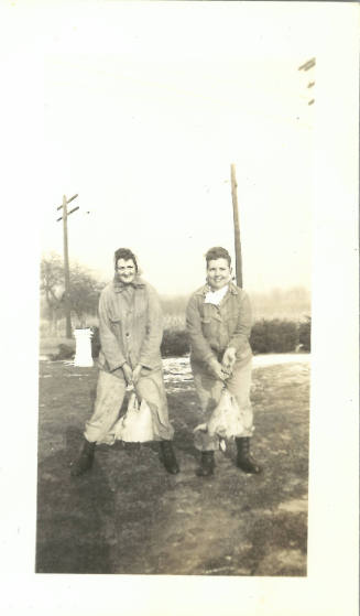 Two women with light skin tone wearing jackets, work boots, and headscarves and holding pigs’ heads