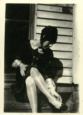 Woman wearing dress with collar sits on porch steps with head lowered as if inspecting her shoe