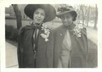 Two smiling women with medium and dark skin tones wear capes, hats, and boutonnieres