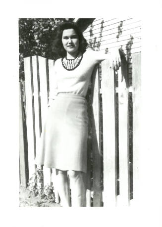 Woman with light skin tone and dark hair wearing skirt poses with arm propped up on fence behind her