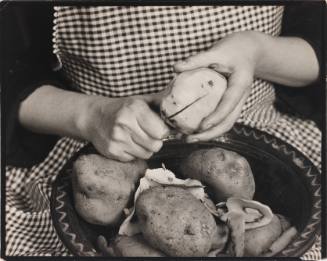 Black-and-white close-up photo of person wearing apron peeling potatoes into a basket on their lap