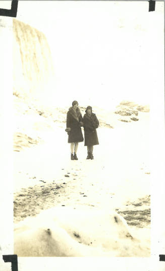 Two women in coats and hats stand facing the camera, a large waterfall on the left behind them