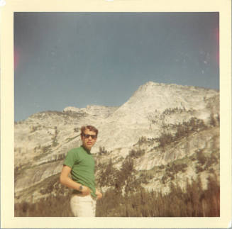 Young man with light skin tone in sunglasses and green t-shirt in front of snow-covered mountains