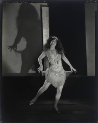 Full-length, dramatically lit portrait of a smiling Ann Pennington posing in fringed stage costume
