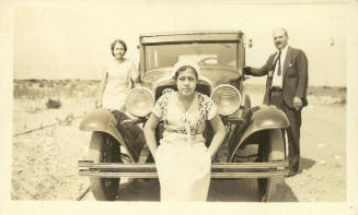 Young woman sits on front bumper of a car; behind her a woman and man stand beside the car