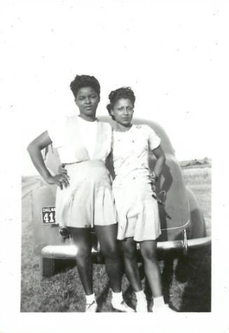 Two young people with dark skin tone wearing light clothes and lace-up shoes stand in front of a car