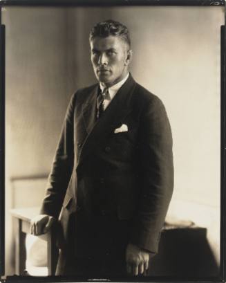 Portrait of Gene Tunney standing with hand on table, wearing dark suit, and looking at camera