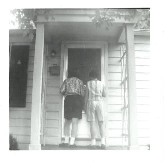 Two people with light skin tone stand on porch of house at the front door, facing away from camera